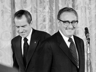 Two men in suits, one behind microphone.