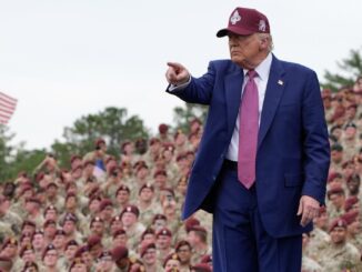 Man in suit pointing, addressing military audience.