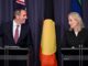 Two people speaking at podiums, flags behind.