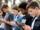Teens focused on smartphones outdoors in line.