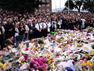Crowd gathers around memorial with flowers.