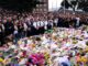 Crowd gathers around memorial with flowers.