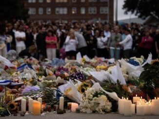 Candlelit memorial with flowers and crowd gathered.