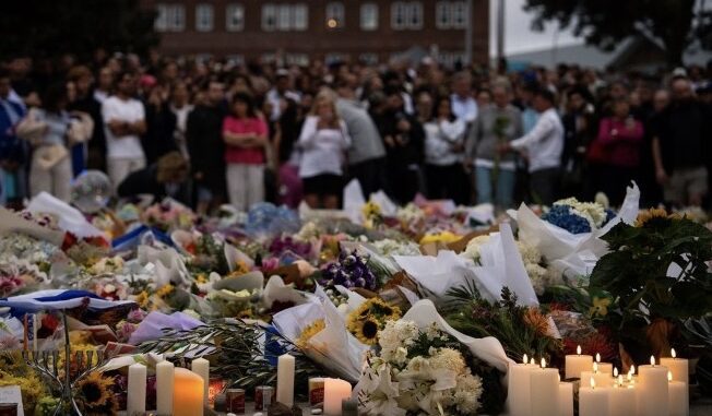 Candlelit memorial with flowers and crowd gathered.