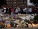 Candlelit memorial with flowers and crowd gathered.