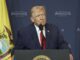 Older man at podium with yellow flag backdrop