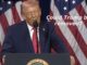 Politician speaking at podium, flags behind.