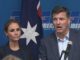 Two people speaking at press conference with flag