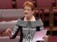 Woman speaking in parliamentary chamber holding paper
