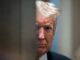 Man with white hair peering through narrow gap